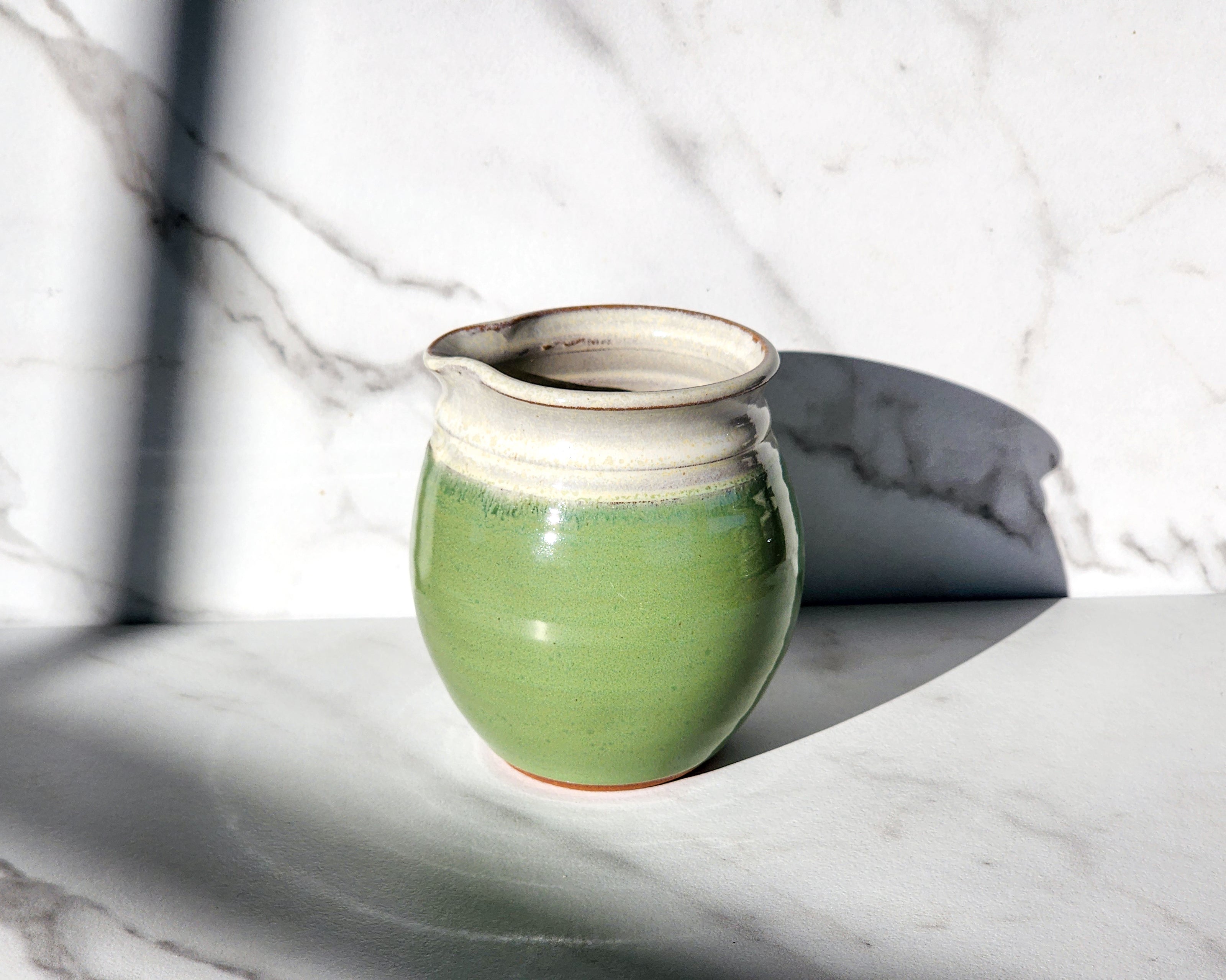 Image Description for Bud Green Creamer: A bud green creamer from Clinton Pottery, featuring a soft, earthy hue reminiscent of freshly sprouted leaves. The creamer's petite size and curved handle offer a charming aesthetic, perfect for enhancing your morning coffee or afternoon tea. Crafted with care and finished with a smooth glaze, this creamer adds a touch of rustic elegance to any tabletop setting.