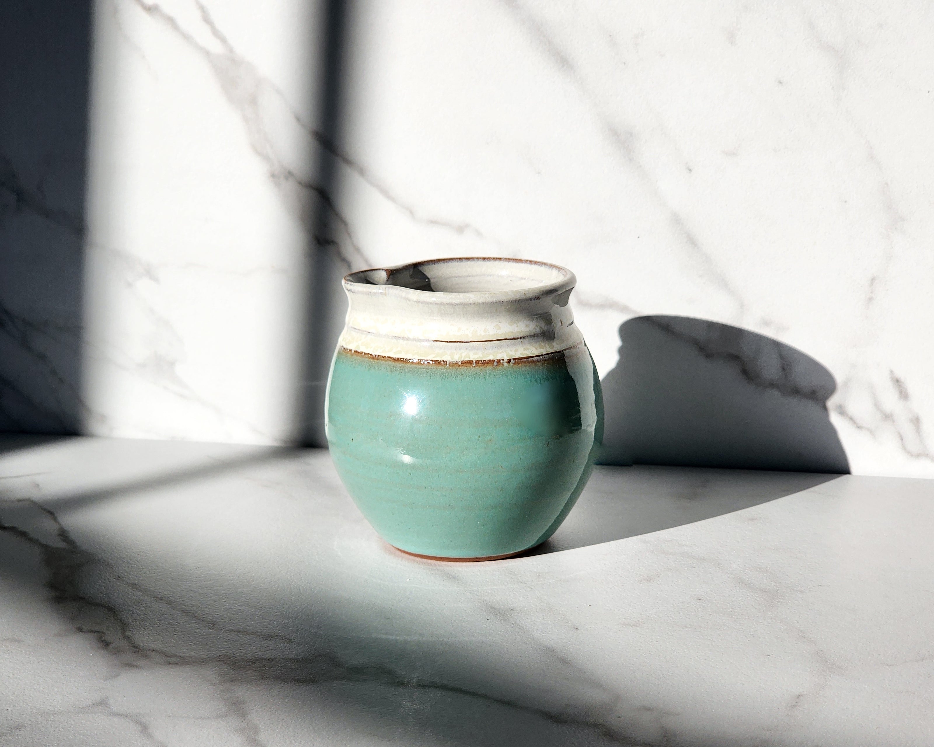 Image Description: A Sky-colored creamer from Clinton Pottery graces a table, its serene hue reminiscent of a clear summer sky. With its smooth surface and graceful curves, this creamer adds a touch of tranquility to any dining setting. Ideal for serving cream or milk alongside coffee or tea, it elevates the dining experience with its subtle yet captivating color..