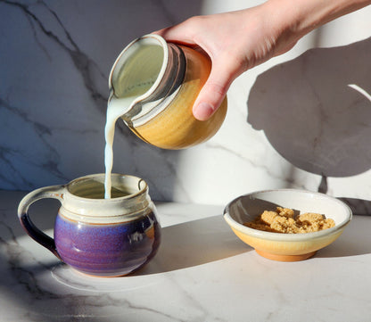  Image Description: A bumblebee yellow creamer pours cream into a small purple mug next to a matching sugar bowl. These vibrant pieces from Clinton Pottery's collection create a charming tableau, adding sunny sophistication to your coffee or tea service.