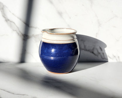  Image Description: A cobalt blue creamer, part of Clinton Pottery's collection, stands elegantly on a table. Its deep blue hue radiates richness and sophistication, adding a pop of color to any tabletop. With its sleek design and glossy finish, this cobalt blue creamer is perfect for serving cream or milk alongside coffee or tea, enhancing your dining experience with a touch of timeless elegance