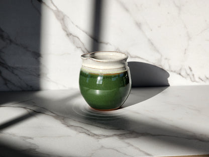 Image Description: A Dark Green creamer by Clinton Pottery sits gracefully on a table, its deep, rich hue evoking the tranquility of a forest canopy. With its smooth surface and sleek design, this creamer adds a touch of natural elegance to any dining setting. Ideal for serving cream or milk alongside coffee or tea.