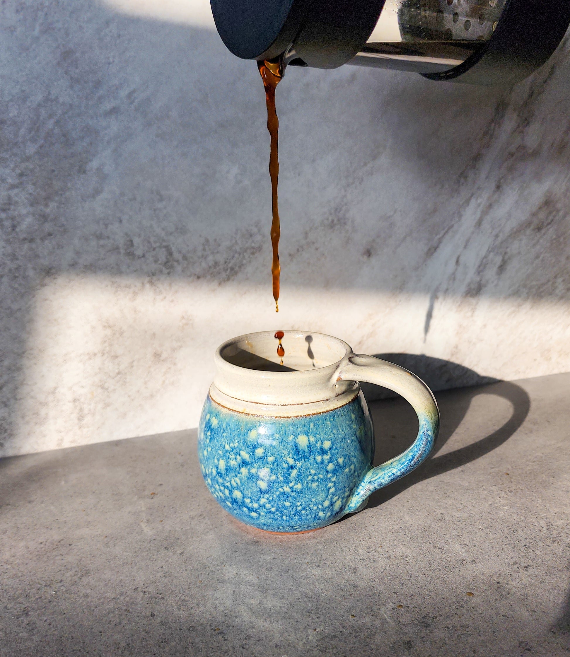  Image: A mesmerizing scene of Clinton Pottery's Handmade Medium Mug in Starry Night capturing the essence of celestial wonder. As coffee gracefully pours into the mug from a French press, the deep, starry blue hue comes to life, enhancing the enchanting appeal of this 14-16 oz masterpiece. Expertly crafted and machine washable for convenience, it seamlessly combines artistic aesthetics with practicality, offering a captivating addition to your daily coffee ritual.